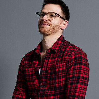 Muž má na sobě kostičkovanou červenou košili a brýle. Usmívá se a stojí před šedým pozadím. | © Andreas Kusy