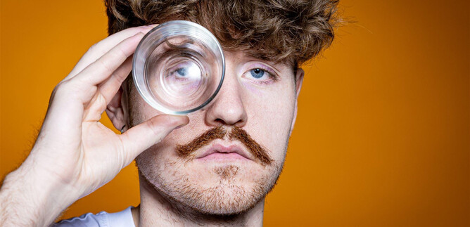 A man with curly hair holds a glass in front of his eye. The background is orange, which gives the image a lively appearance. | © Anna Sophie Kölbl