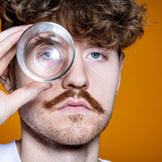 Ein Mann mit lockigem Haar hält ein Glas vor sein Auge. Der Hintergrund ist orange, was das Bild lebhaft wirken lässt. | © Anna Sophie Kölbl