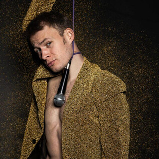 Mladý muž má na sobě třpytivý oblek a drží mikrofon v ruce. Pozadí je pokryto zlatým třpytem. | © Yuting Tseng