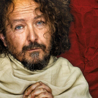 Egy férfi göndör, barna hajjal és szakállal gondolkodó arckifejezéssel néz a kamerába. Egyszerű, világos ruhát visel egy piros háttér előtt. | © Bernhard Eder