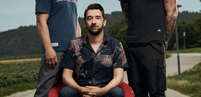 Three men are standing on a street. The man in the middle is sitting on a red chair, while the other two are standing and posing in fashionable clothes. | © Pascal Riesinger