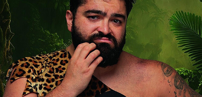A man in a leopard skin with a thoughtful expression. In the background, green plants and jungle can be seen. | © Serdar Karibik