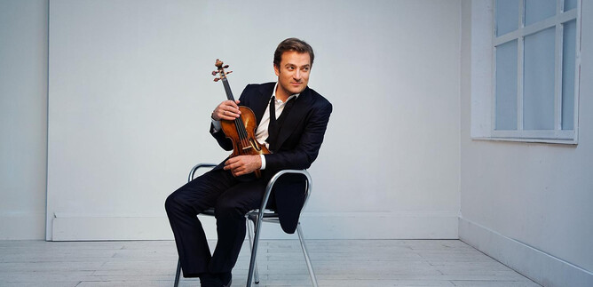 A man is sitting on a chair and holding a violin. The background is simple and bright, which draws the focus to him. | © Simon Fowler