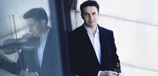 Ein Mann mit einer Geige steht vor einer reflektierenden Oberfläche. Er trägt einen schwarzen Anzug und lächelt freundlich. | © Nikolaj Lund