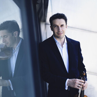 Muž s houslemi stojí před reflexním povrchem. Má na sobě černý oblek a přátelsky se usmívá. | © Nikolaj Lund