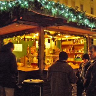 Ein festlich beleuchteter Stand auf einem Weihnachtsmarkt. Menschen stehen zusammen und genießen die Atmosphäre und das Angebot. | © LionsClub