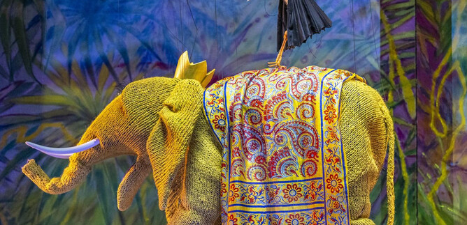 Ein bunter Elefant mit einem prächtigen Sattelmuster steht im Vordergrund. Auf seinem Rücken balanciert ein Storch. | © Bernhard Müller