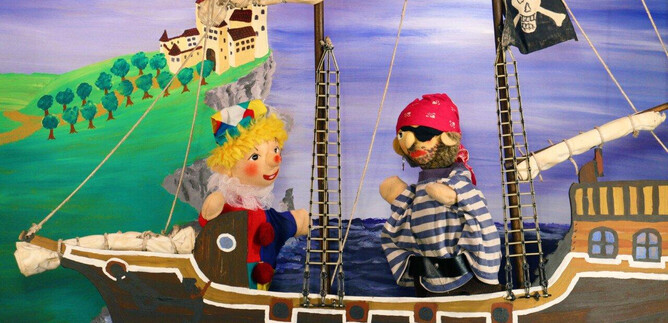 A puppet theater on a ship with a pirate and a happy girl. The backdrop features a green landscape with a castle in the background. | © Friedburger Puppenbühne
