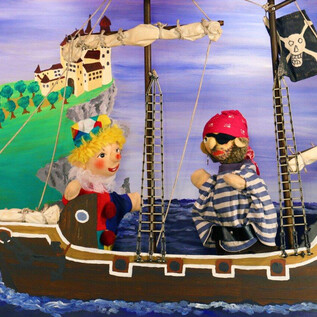 Un teatro delle marionette su una nave con un pirata e una ragazza felice. Lo sfondo mostra un paesaggio verde con un castello sullo sfondo. | © Friedburger Puppenbühne