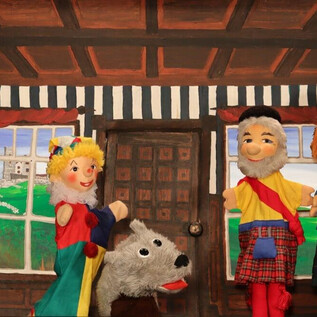 Drei Handpuppen stehen in einem stilvollen Raum mit Holzvertäfelung. Ein Hund aus Fell liegt vor ihnen, während Gemälde an den Wänden hängen. | © Friedburger Puppenbühne