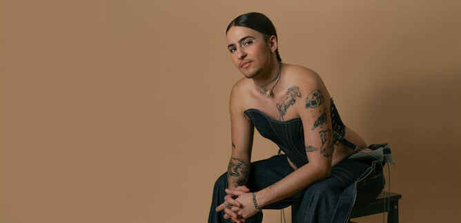 Eine Person sitzt auf einem Stuhl vor einem beigen Hintergrund. Sie trägt ein modisches Outfit und hat sichtbare Tattoos. | © Sudimac Ivana