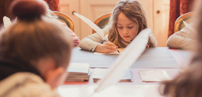 A girl is writing at a table, surrounded by other children. On the table are a pen, paper, and school supplies. | © Eva trifft