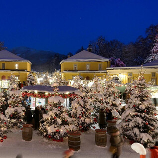 A festive Christmas market at night, surrounded by snow-covered trees and atmospheric lights. Visitors stroll between festively decorated stalls. | © Kinderchor Unison