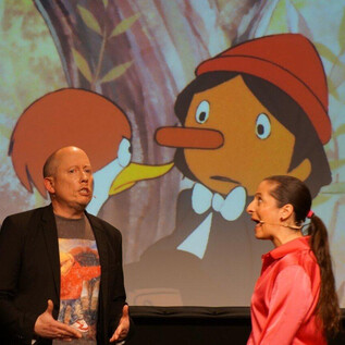Una actuación en vivo con dos intérpretes en el escenario. Al fondo se puede ver una escena de una película de animación. | © Sole Goscinski