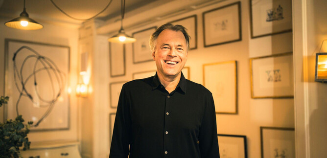 Un hombre sonriente está de pie en una habitación elegante con paredes iluminadas. En el fondo, hay cuadros enmarcados que crean una atmósfera creativa. | © Mina Esfandiari
