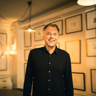 A smiling man stands in a stylish room with illuminated walls. Framed pictures hang in the background, creating a creative atmosphere. | © Mina Esfandiari