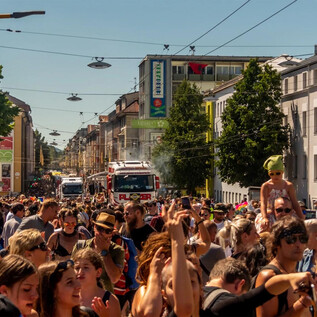 Une rue animée avec une grande foule qui fête un jour ensoleillé. À l'arrière-plan, on peut voir des arbres et des bâtiments, ainsi qu'un camion de pompiers. | © www.uniteparade.at