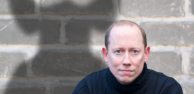 Un uomo è seduto di fronte a un muro grigio con un'espressione seria. Sullo sfondo è visibile un'ombra che ha una forma interessante. | © ARGEkultur