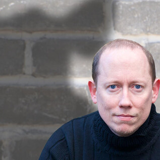 Ein Mann sitzt vor einer grauen Mauer mit einem ernsten Ausdruck. Im Hintergrund ist ein Schatten sichtbar, der eine interessante Form hat. | © ARGEkultur