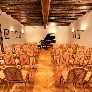 Un espacio elegante con suelo de madera y vigas en el techo. En el centro hay un piano, rodeado de muchas sillas para el público. | © Agentur Orpheus