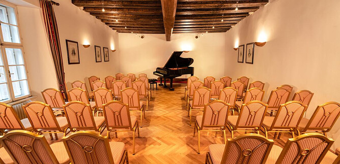 An elegant room with wooden floors and ceiling beams. In the center stands a piano, surrounded by many chairs for the audience. | © Agentur Orpheus