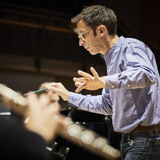 A conductor leads an orchestra with passionate hand gestures. In the foreground, a flute is visible. | © Christophe Raynaud