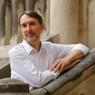 A man in a white shirt is leaning casually against a staircase. In the background, parts of historic buildings can be seen. | © Deyan Parouchev