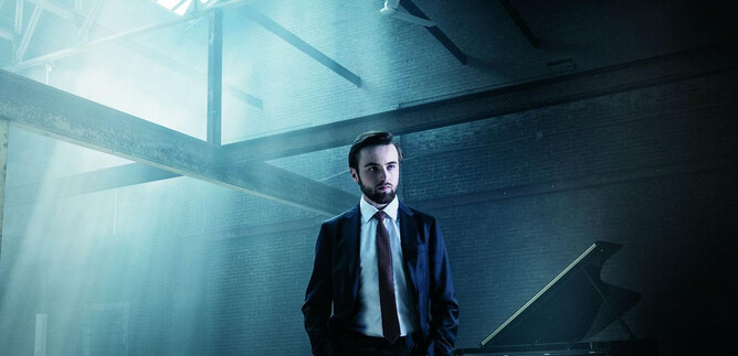 Egy öltönyös férfi áll egy világos, fényben gazdag szobában. A háttérben egy zongora áll. | © Dario Acosta