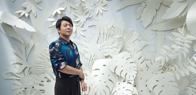 A man stands in front of an ornate white wall with plant motifs. He is wearing a colorful shirt and holding a flower in his hand. | © Olaf Heine