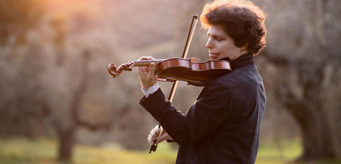 A young man is playing the violin in a tranquil landscape. In the background, trees and soft light can be seen. | © Suxiao Yang