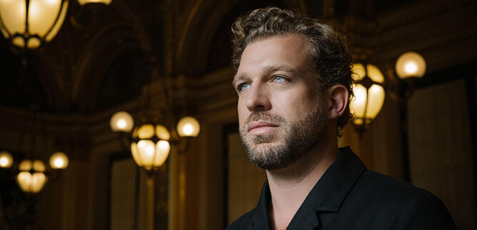 A man with curly hair stands in an elegant room. The background features historical lamps and stylish architecture. | © Chris Gonz