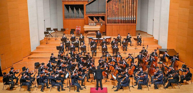 Une grande orchestre joue sur une scène. Le chef d'orchestre dirige les musiciens, qui sont vêtus de costumes élégants. | © Konzert Meiji University Nakano