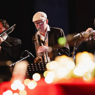 Egy zenekar játszik a színpadon, miközben a zenészek trombitál és szaxofonoznak. A háttérben lágy fények láthatók, amelyek ünnepi hangulatot teremtenek. | © Stefan Rochhart