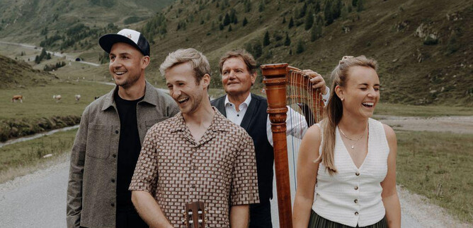 A group of four people is standing on a road in a picturesque landscape. They are smiling and posing, while gentle hills can be seen in the background. | © Bergstadtmomente