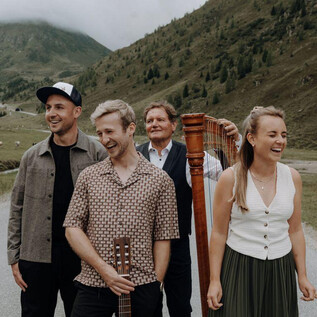 A group of four people is standing on a road in a picturesque landscape. They are smiling and posing, while gentle hills can be seen in the background. | © Bergstadtmomente