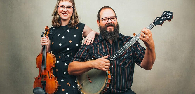 Ein Mann und eine Frau posieren zusammen mit Instrumenten. Die Frau hält eine Geige und der Mann spielt Banjo, beide lächeln in die Kamera. | © Sandlin Gaither