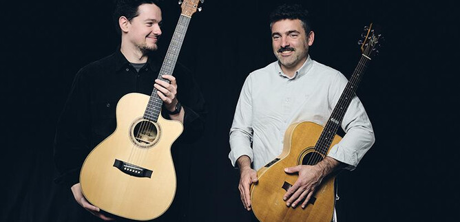 Zwei Männer stehen lächelnd nebeneinander und halten Akustikgitarren. Sie wirken freundlich und entspannt vor einem dunklen Hintergrund. | © Elias Maier