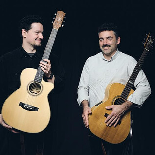 Two men are standing side by side, smiling, holding acoustic guitars. They appear friendly and relaxed against a dark background. | © Elias Maier