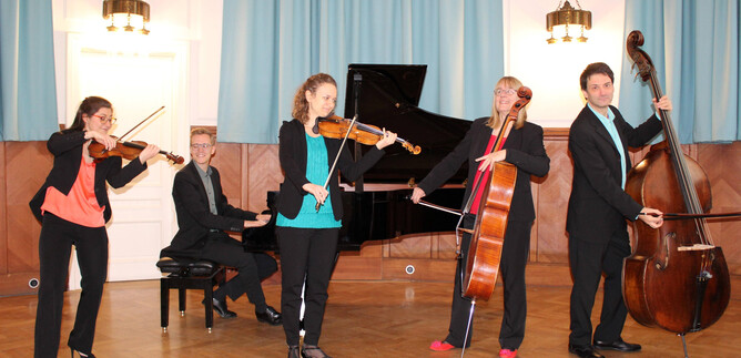 A music group is playing in an elegant room. Four musicians with different instruments are standing together, while a pianist is sitting at the piano. | © Ehrbarsaal