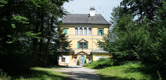 A large, yellow house surrounded by green trees. The path leads directly to the entrance of the building. | © Salzburg Museum