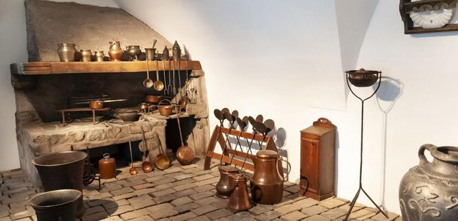 A historic kitchen with old cooking utensils and a stone fireplace. The floor is made of large stones, and there are various pots and jugs. | © Salzburg Museum