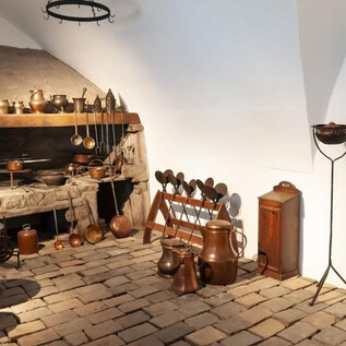 A historic kitchen with old cooking utensils and a stone fireplace. The floor is made of large stones, and there are various pots and jugs. | © Salzburg Museum