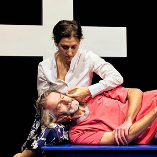 Une femme est assise à côté d'un homme qui est allongé sur un banc. En arrière-plan, une grande croix blanche est visible. | © Chris Rogl