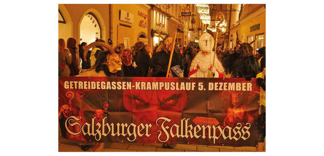 Egy tömeg részt vesz egy fesztiváli felvonuláson, miközben egy nagy transzparenst tart, amin a „GETREIDEGASSEN-KRAMPUSLAUF 5. DECEMBER” felirat áll. Elöl láthatók öltönyökbe öltözött emberek, akik hagyományos figurákat ábrázolnak. | © Salzburger Falkenpass