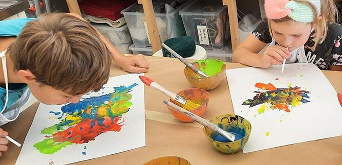 Két gyerek ül egy asztalnál, és színes festékekkel festenek. Előttük festékkel teli tálak és a papíron készült műveik állnak. | © DomQuartier