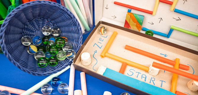 Colorful crafting materials are spread out on a wooden table. There are pieces of paper, scissors, and craft sticks in various colors visible. | © Salzburg Museum/Maurice Rigaud