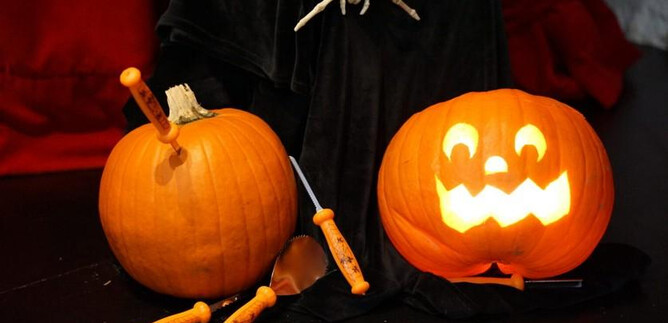 Dos calabazas con motivos de Halloween están sobre una mesa. Una calabaza es luminosa y tiene una cara alegre, mientras que la otra está decorada con herramientas. | © Salzburg Museum