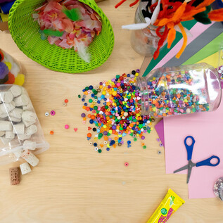 A colorful crafting station with various materials such as beads, colorful paper, and scissors. Pom-poms and other crafting supplies are also spread out on the table. | © Salzburg Museum/Maurice Rigaud