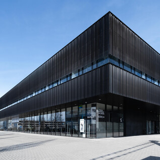 Two hikers are traversing a green, hilly landscape with some rocks and a calm lake in the background. The sky is cloudy, and the scenery appears wild and untouched. | © Messezentrum Salzburg/Sarah Fellner